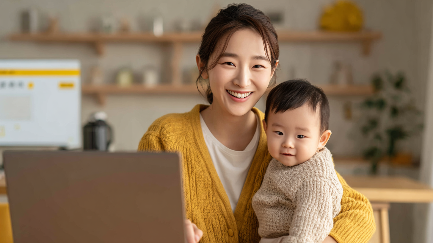 아기를 안은 웃는 한국 어머니가 노트북으로 아동수당 온라인 신청을 하는 모습