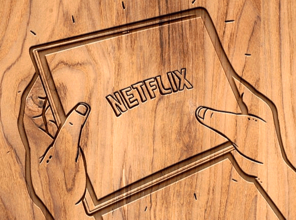 나무판에 남자의 두 손이 새겨져 있고 두 손은 사각형의 태블릿을 들고 있다. 태블릿 안에는 영어로 넷플릭스가 적혀있다.