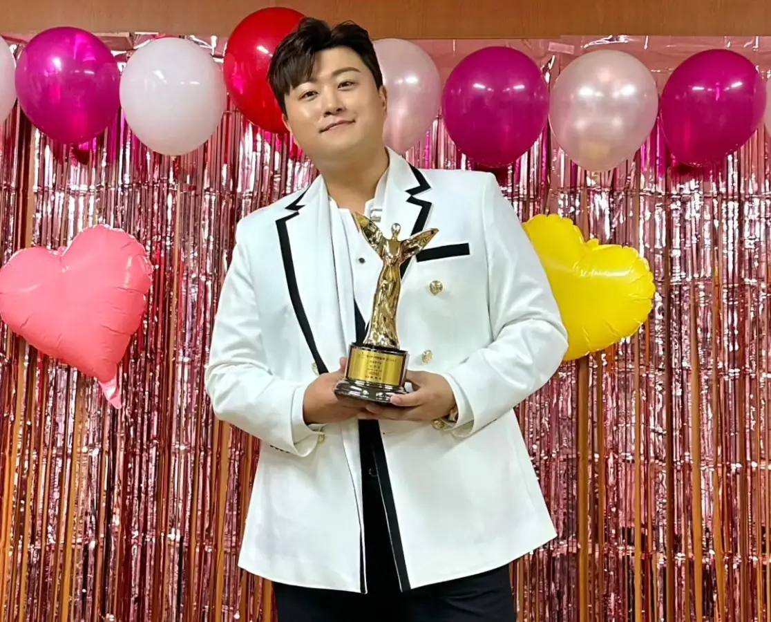 분홍색 반짝이와 풍선들을 배경으로 흰색 쟈켓을 입은 김호중이 두손으로 황금색 트로피를 들고 미소짓는 모습