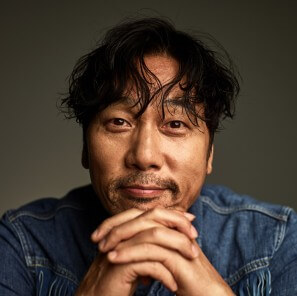 이문세가-청자켓을-입고-양손을-깍지-낀-채-턱을-괴고-살짝-미소를-띄고-있는-사진으로-눈은-슬퍼보인다.