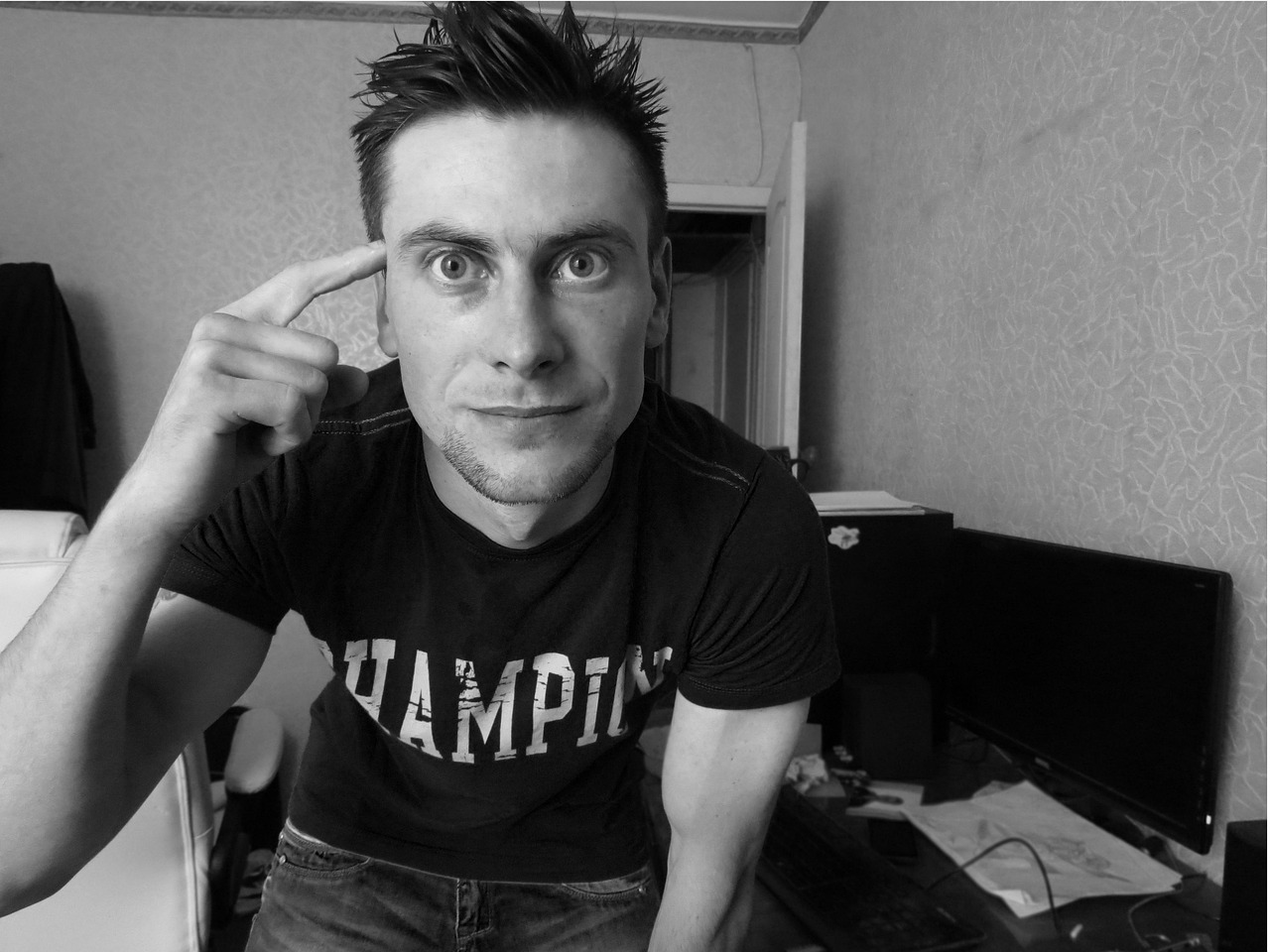 흑백 톤으로 촬영된 실내 인물 사진으로, 프레임 중앙에는 짧은 헤어스타일과 진지한 눈빛의 남성이 등장한다. 그는 "CHAMPION"이라는 글자가 적힌 반팔 티셔츠를 입고 있으며, 한 손가락을 자신의 관자놀이에 갖다 대는 포즈를 취하고 있어 생각 혹은 아이디어를 상징하는 제스처로 보인다. 배경에는 열려 있는 방문과 어지럽혀진 책상, 모니터, 문서 등이 놓여 있으며, 전반적으로 개인 작업 공간 혹은 방 안의 자연스러운 분위기를 드러낸다. 구도는 광각 렌즈로 가까이 다가간 시점에서 촬영되어 인물의 표정과 자세가 과장되게 표현되며, 강한 몰입감을 유도하는 사진.