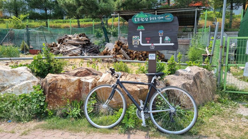 등산로 안내판 기둥에 기대어 매어놓은 자전거, 뒤는 텃밭, 그린철망,