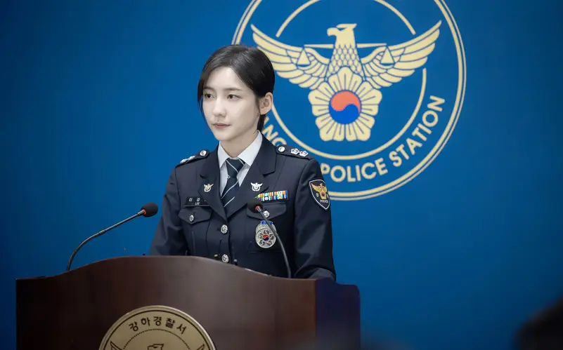 재벌X형사- 파란배경 가운데 노란색 독수리 경찰관마크가 그려진 벽면앞 갈색 단상 위 마이크 2개앞에서 경찰제복을 입고 서있는 이강현