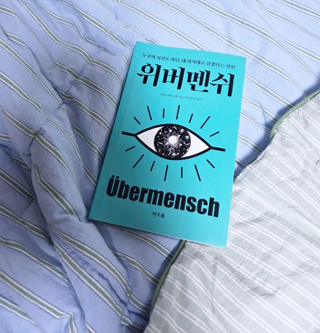 자기 전 침대 위에 놓인 위버멘쉬 책 표지, 청록색 바탕에 우주가 담긴 눈동자 일러스트