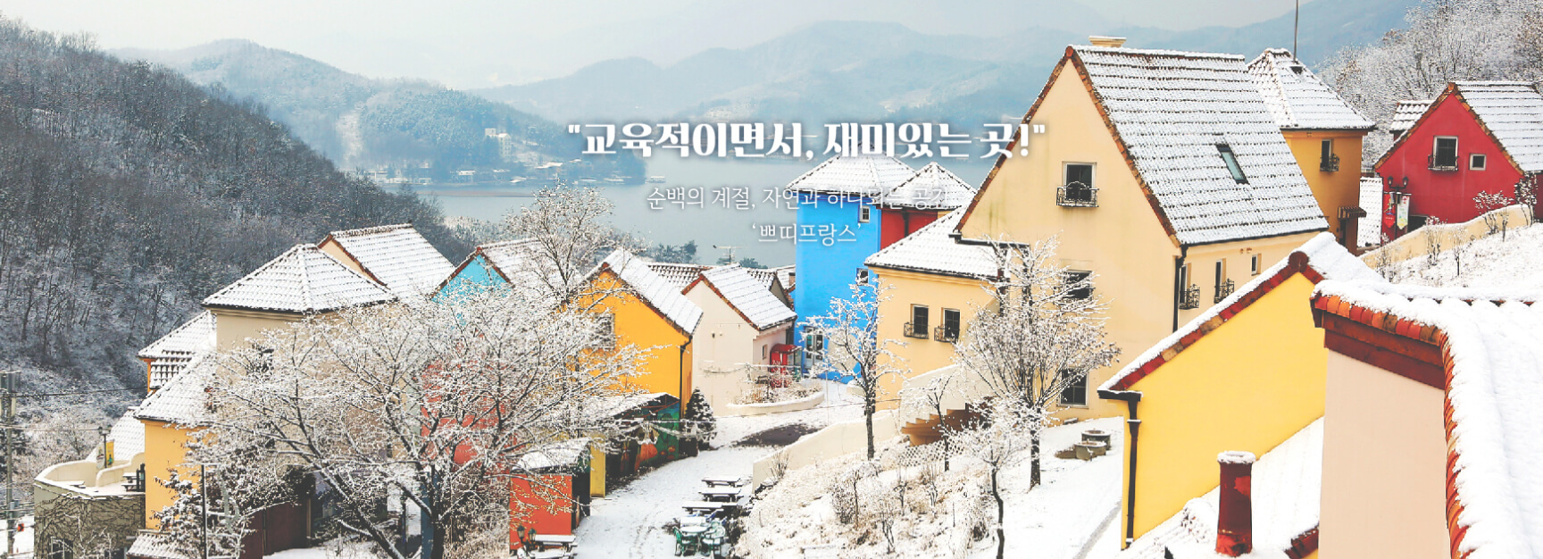 눈 덮인 아름다운 마을, 쁘띠프랑스의 전경. 알록달록한 건물들이 순백의 눈과 어우러져 동화 같은 분위기를 자아내며, 뒤편에는 조용히 흐르는 강과 설산이 배경으로 펼쳐져 있습니다