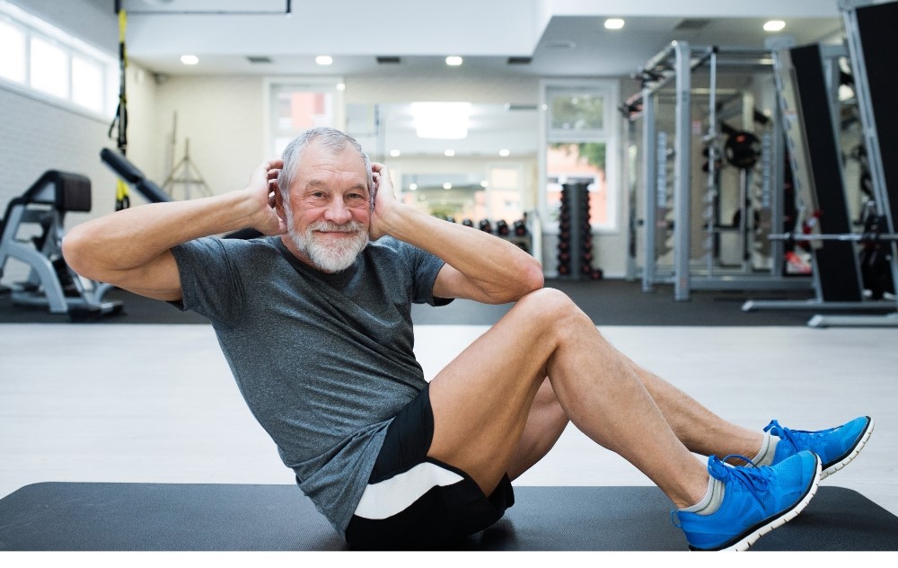 건강한 중년 남성이 헬스장에서 운동하는 모습, 회색 운동복을 입고 윗몸일으키기를 하며 밝게 웃고 있다 via canva