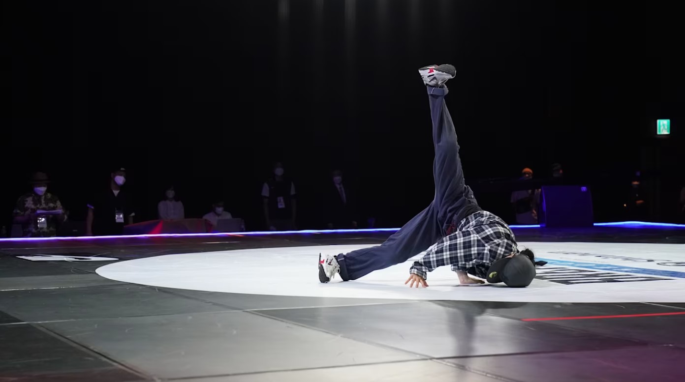 한 남성이 경기장 중앙에서 화려한 브레이킹 댄스를 선보이며 열정적인 경연을 펼치고 있습니다. 그의 뒤에는 수많은 관객과 심사위원들이 자리하고 있습니다.