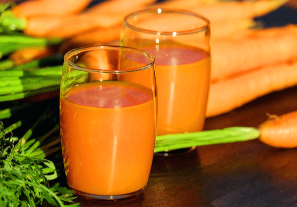 나무 식탁 위에 투병 유리컵에 담긴 주황색 당근주스 두 잔이 있다. 컵 뒤에는 당근들이 가득하다.