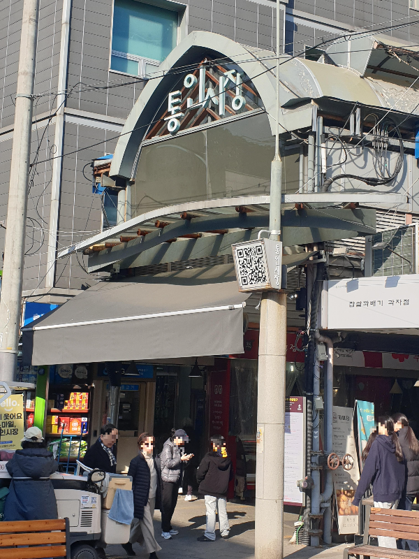 통인시장 출입구 위쪽에 통인시장이라는 간판이 달려있다. 앞에 사람들이 지나다닌다.