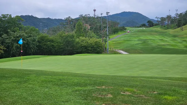 산과 숲을 배경으로 그린의 깃대와 길게 이어진 페어웨이가 보이는 골프장 전경