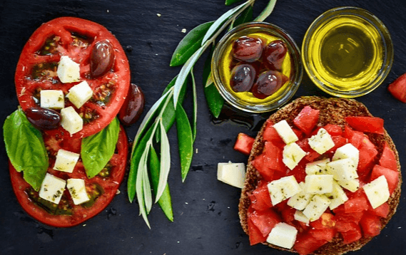 검정 식탁에 얇게 저민 토마토 2개가 있고 그 위에는 올리브와 치즈덩이가 올려졌다. 옆에는 2개의 작은 유리그릇에 담긴 올리브유가 있다.