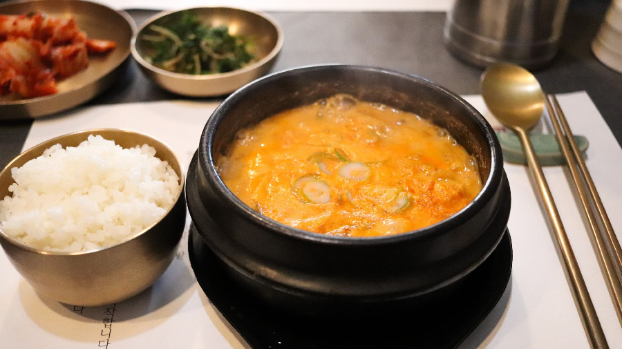 식탁 위에 흰색 종이를 깔고 그 위에 쌀밥과 된장찌개를 담은 뚝배기와 김치, 시금치를 담은 그릇을 놓아두고 숟가락과 젓가락을 옆에 놓아두고 찍은 사진