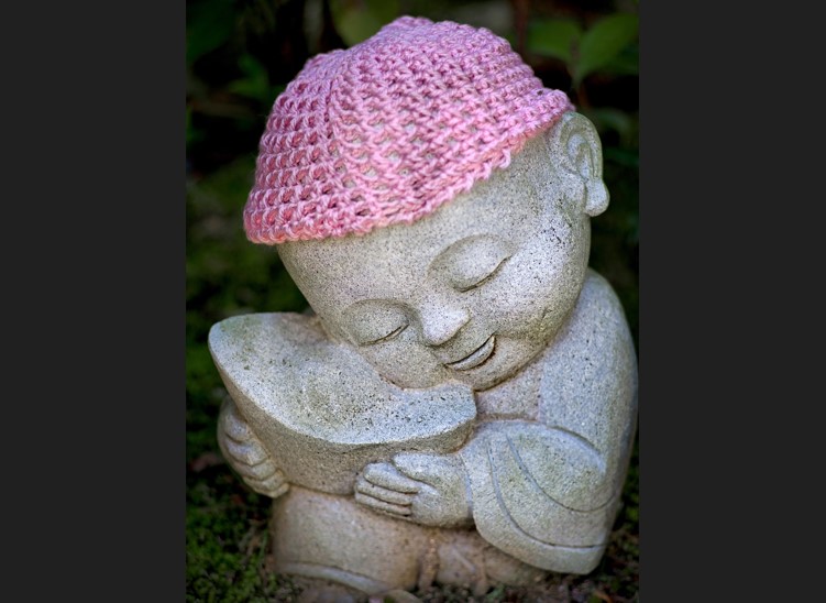 어리고 귀여운 동자승의 모습을 조각한 조각상이 풀밭에 앉아 있는데, 머리에 핑크빛 뜨게 모자가 씌워져 있는 사진