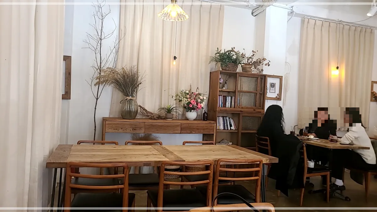 동탄플랜츠카페내부-나무테이블과-의자들-화병의분홍색꽃-은은한간접조명-브라운색의책장과-책들-테이블에서-커피를-마시며-대화를-나누는-3명의-여자들