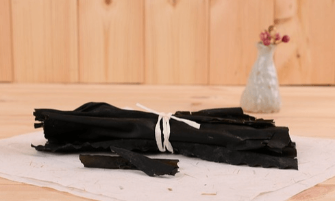 나무 바닥에 하얀 천이 깔려있고 그 위에 검정색의 마른 다시마 한뭉치가 놓여있다.