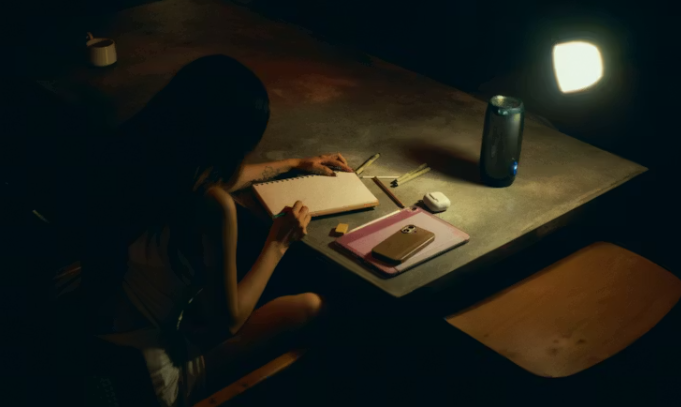 따뜻한 조명이 켜진 어두운 방에서 한 여성이 노트에 펜으로 무언가를 쓰고 있다. 테이블 위에는 다이어리, 스마트폰, 펜, 이어폰 등이 놓여 있고, 조용하고 사적인 분위기가 감돈다.