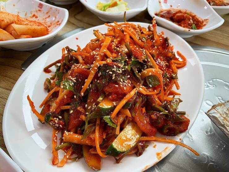 오이, 상추 등 야채와 서대회가 빨란 양념에 무쳐져서 하얀 접시 위에 놓여 있다. 음식 위에서 깨가 뿌려지 있다.