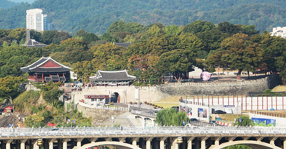Jinjuseong’s stone walls, tiled gate and riverside pavilion stand beneath trees, with a bridge across the foreground.