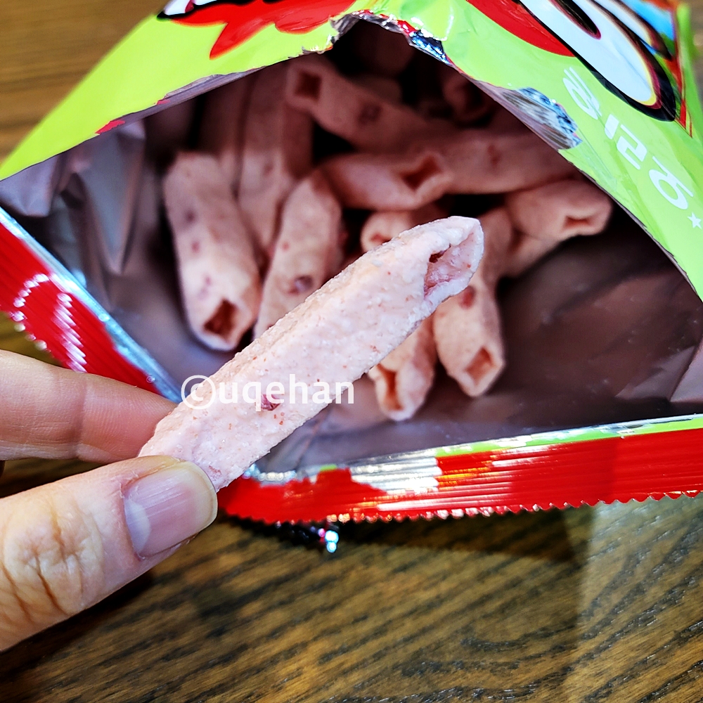 오리온 오! 딸기 과자를 손으로 잡고 있따. 긴 막대기에 가운데가 뚫려있따. 과자 중간에 분홍색 딸기 쿠키가 부서져 붙어있다.