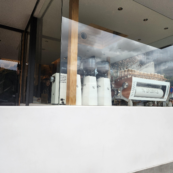 흰 벽에 넓은 창. 그 창에 그라인더와 커피 머신이 보인다. 모든 머신은 흰색으로 구성되어 있다.