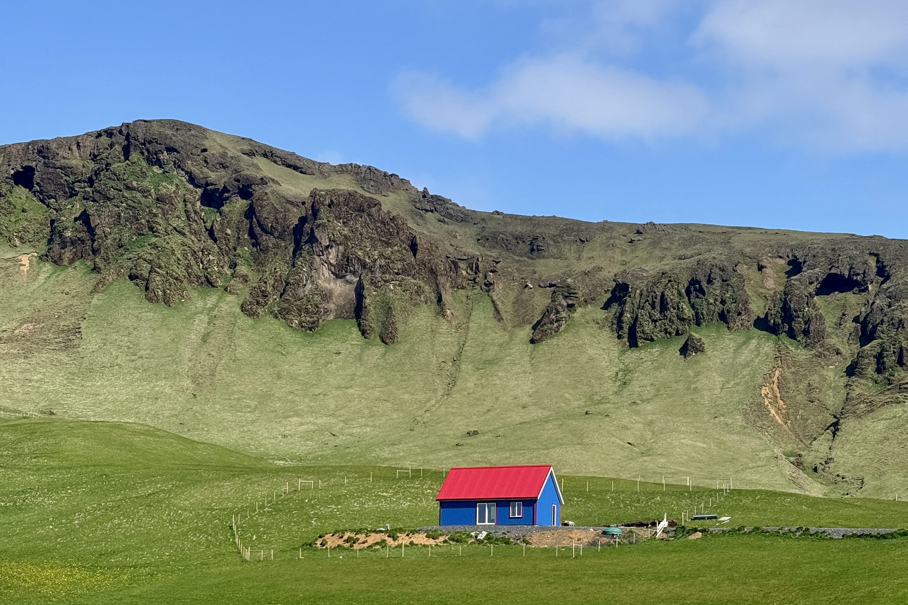 초록 언덕과 산 배경 앞에 있는 빨간 지붕의 파란 집. 파란색 벽에 흰색 창문이 있는 작은 단독주택으로, 주변은 울타리로 둘러싸인 초원 지대임.