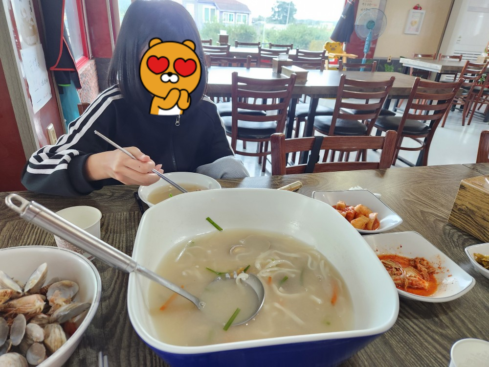 커다란 그릇에 바지락이 가득 담긴 해물칼국수. 아이가 국자로 칼국수를 뜨며 맛있게 먹고 있다.