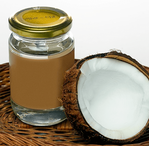 투명 유리병에 코코넛오일이 가득하고, 옆에는 반이 잘린 하얀 속살과 갈색 껍질을 가진 코코넛이 있다.