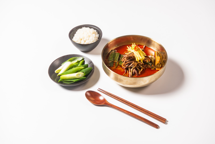 하얀 바닥 위에 검고 동그란 밥그릇에 쌀밥을 가득 담아두고&#44; 그 밑에는 검고 동그란 접시에 파를 썰어 놓은 것을 담아 두고&#44; 그 옆에는 금색 동그란 그릇에 육개장을 가득 담아두고&#44; 그 밑에는 나무 숟가락과 젓가락을 두고 찍은 사진