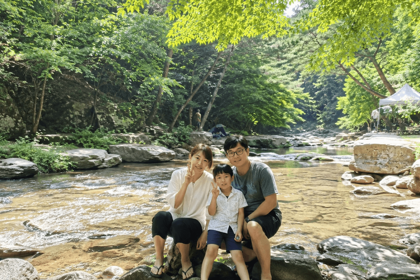 울창한 숲으로 둘러싸인 수태골 계곡의 맑은 물이 흐르는 바위 위에서 방문객들이 앉아 있는 모습, 뒤편으로 텐트와 숲이 보이는 여름 풍경