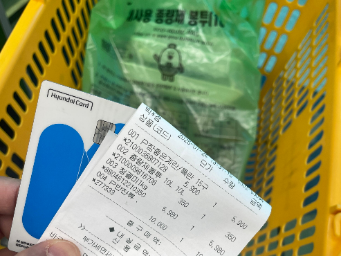 노란 장바구니 안에 쓰레기 봉투와 그 안에는 구입한 물건들이 담겨져 있고, 바구니 위쪽으로 카드와 영수증을 들고 있는 손이 있는 사진입니다.