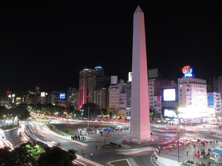 밤의 아르헨티나 부에노스아이레스 시내 전경. 중앙에 거대한 하얀색 오벨리스크가 우뚝 솟아 있고, 그 아래 넓은 7월 9일 대로 위로 자동차들이 지나가며 만들어낸 불빛 궤적이 역동적으로 펼쳐져 있음. 주변에는 화려한 전광판이 달린 현대적인 빌딩들과 가로등 불빛이 어우러진 대도시의 야경.