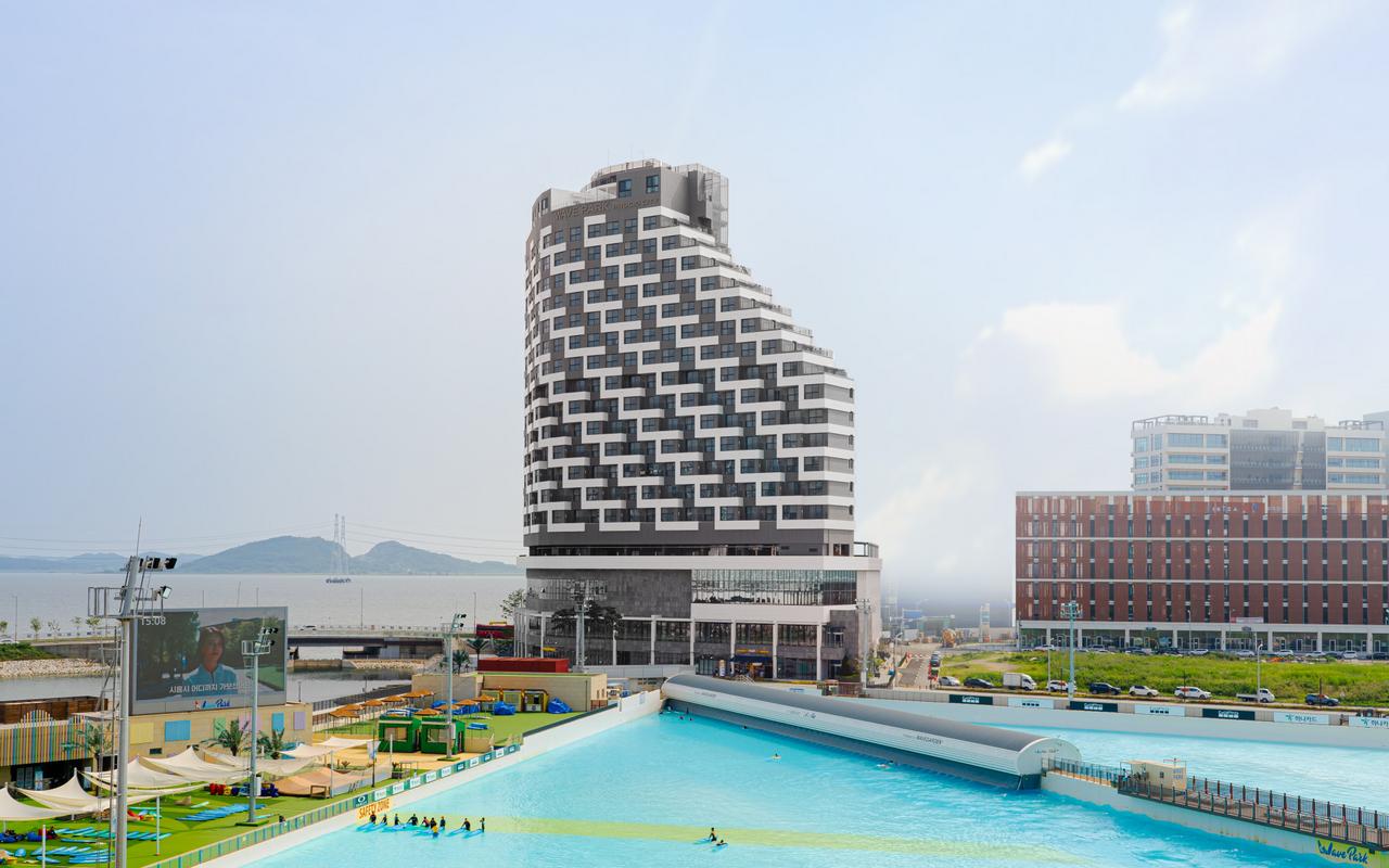 경기도 시흥시 거북섬에 위치한 르 컬렉티브 시흥 웨이브파크 전경. 파도풀과 서핑 시설이 어우러진 워터파크 앞에 독특한 외관의 고층 호텔 건물이 자리해 있으며, 바다와 연결된 탁 트인 전망이 인상적이다.
