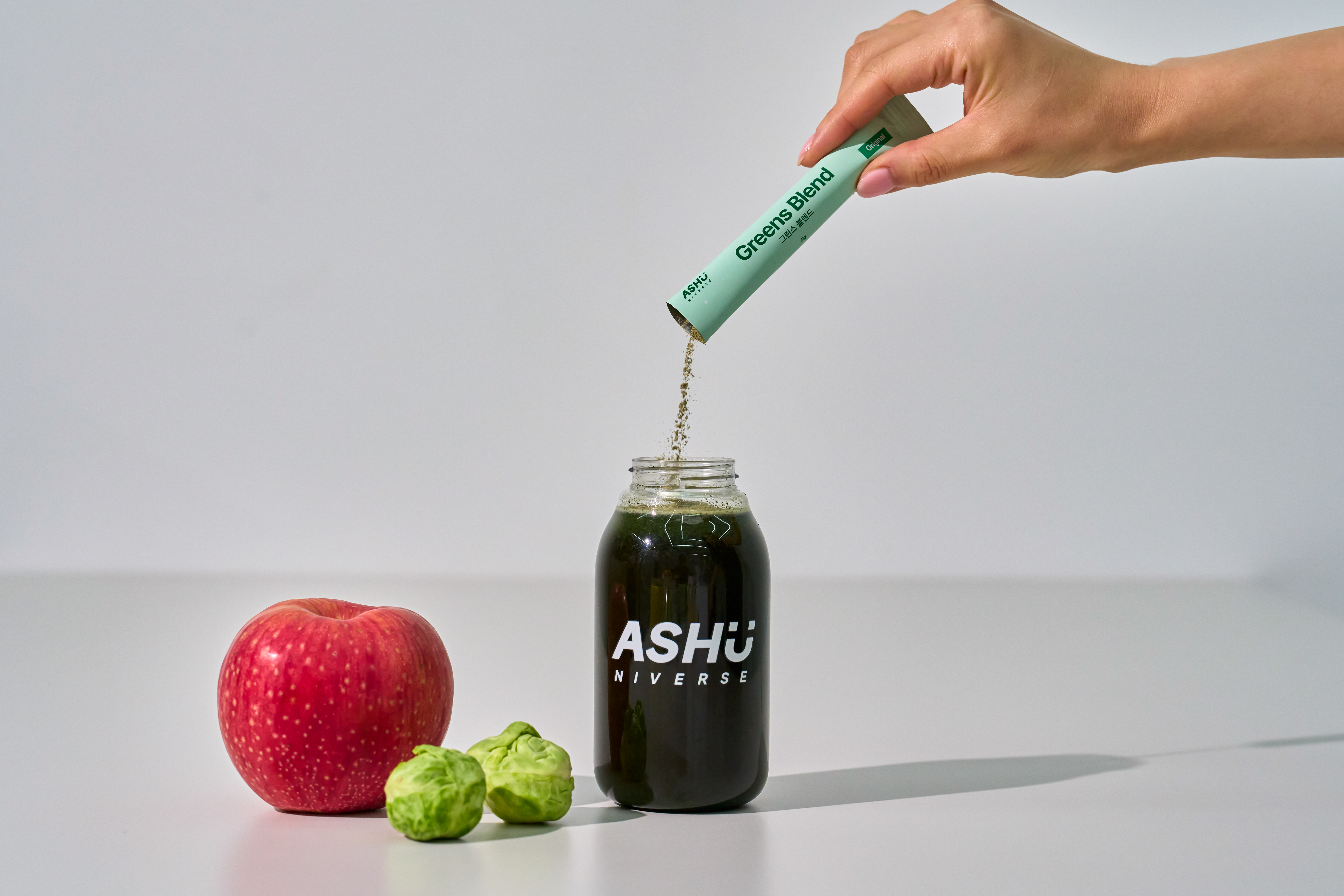 ASHUNIVERSE 그린스블렌드 스틱을 병에 넣어 녹색 음료를 만드는 장면과 사과, 채소 이미지