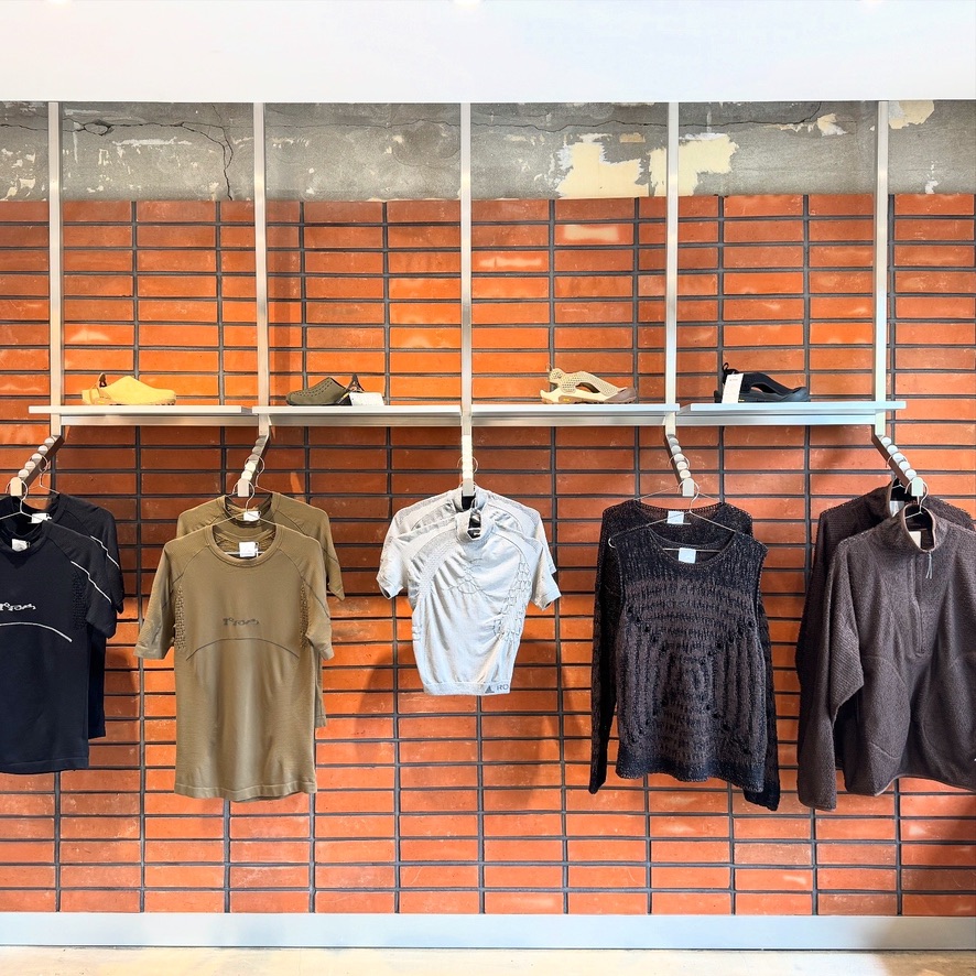 붉은 벽돌 배경 매장에서 티셔츠와 긴팔 상의가 걸려 있고, 상단 선반에는 운동화와 샌들이 전시된 모습