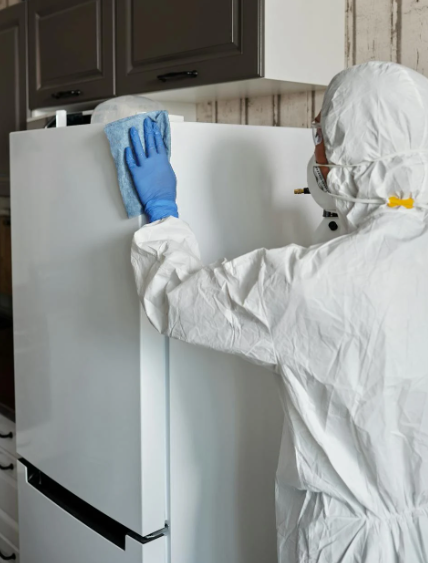 방호복과 고무장갑, 마스크를 착용한 사람이 냉장고를 소독하며 청소하는 모습. 위생을 위한 냉장고 외부 살균 장면.
