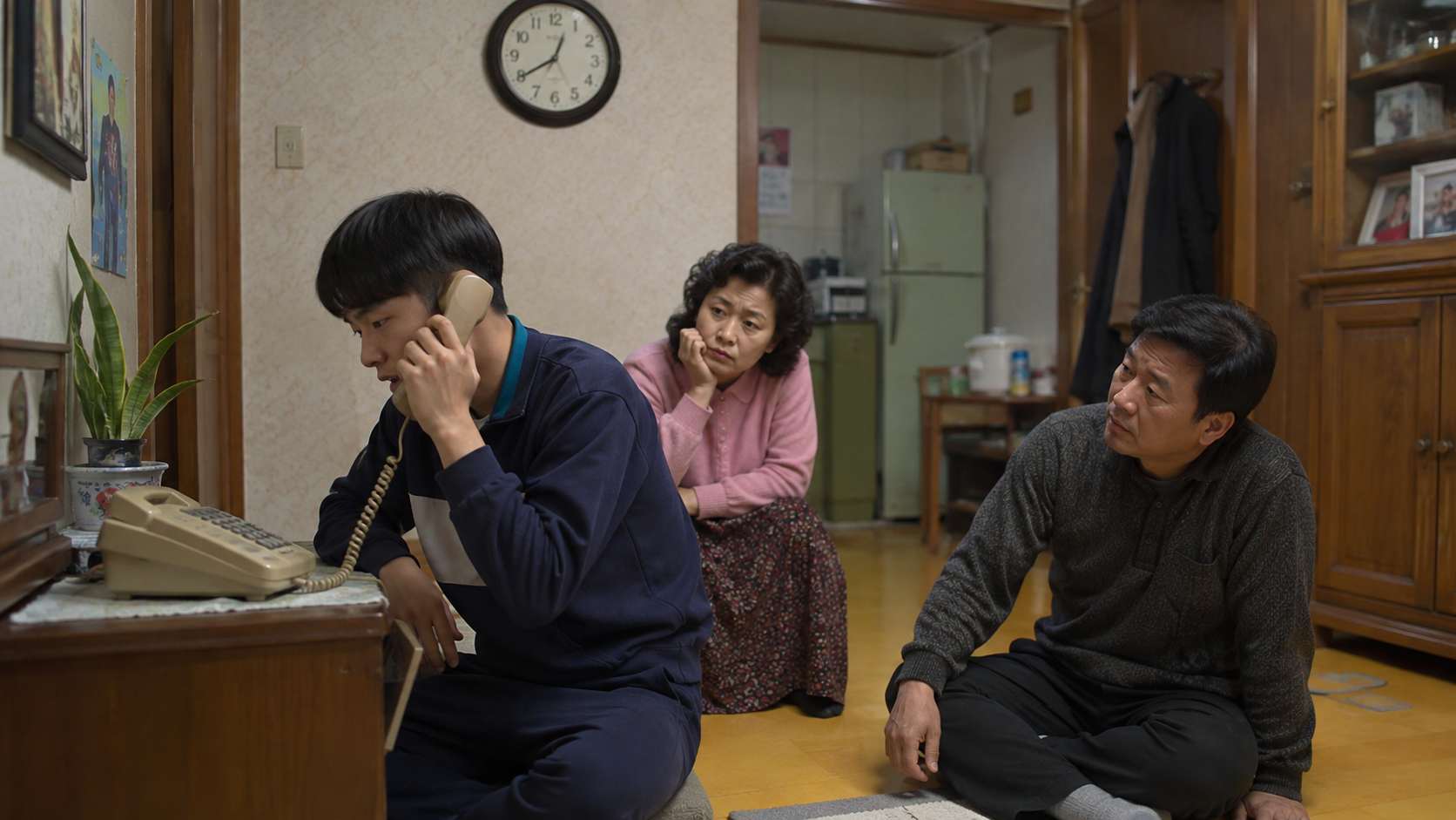 소년이 집 전화로 통화하는 동안 뒤에서 부모님이 앉아 기다리며 바라보는 모습