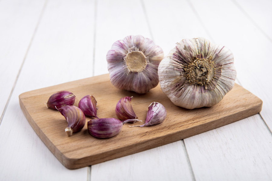 흰색 바닥 위에 나무로 만든 네모난 깔판을 깔고 그 위에 통마늘 2개와 쪼갠 마늘 6개를 놓아두고 찍은 사진