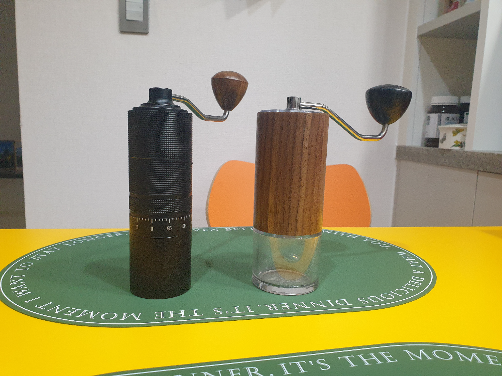 노란 테이블 위에 초록 매트가 깔려 있고 그 위에 검은색 그라인더 하나외 우드 그라인더(우측)이 올려져 있다.