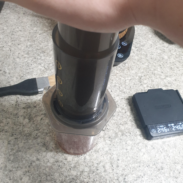 서버 위에 에어로프레스를 손바닥으로 지그시 누르는 모습 바닥에는 붓, 저울, 드립포트가 보인다.