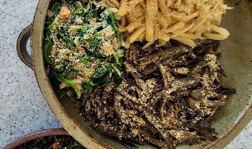 나물, 무나물, 취나물 등 다양한 건강 나물이 담긴 양은 냄비. 나물 위에는 깨가 솔솔 뿌려져 있으며, 전통적인 건강 밥상을 연상케 한다.