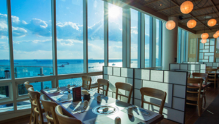 식당 내부에 테이블이 있고 햇빛이 창문으로 들어오고 있다 창밖으로는 바다가 보인다