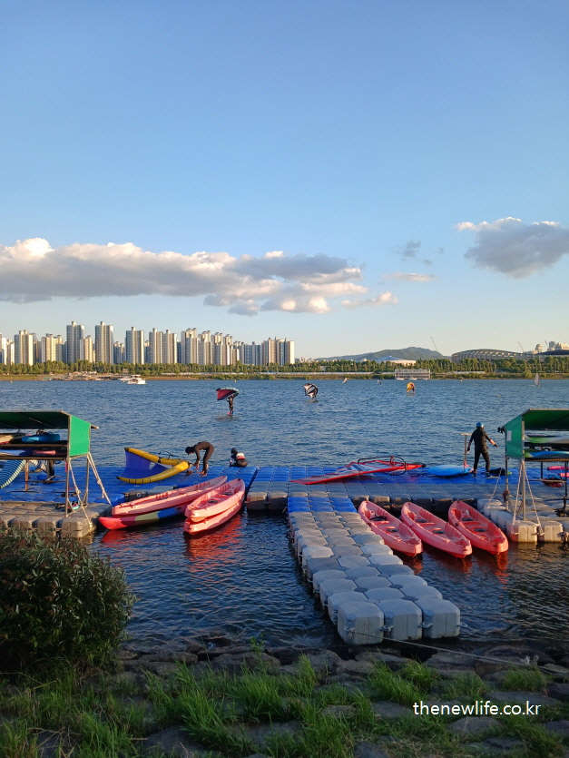 People kayaking and paddleboarding on the Han River with a backdrop of tall apartment buildings./고층 아파트를 배경으로 한강 위에서 카약과 패들보드를 즐기는 사람들.
