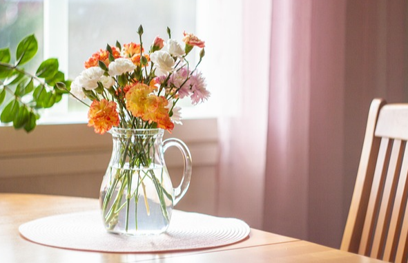 나무 식탁에 투명한 화병이 있다. 그 안에는 주황색과 하얀색 꽃이 가득하다.