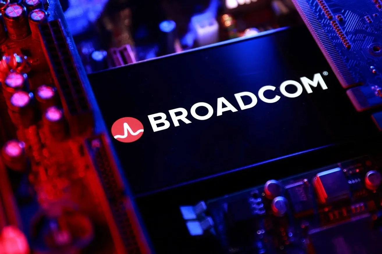 스마트폰 화면에 Broadcom 로고가 크게 표시되어 있다. 로고는 흰색 글씨로 BROADCOM이라고 쓰여 있으며, 왼쪽에는 빨간색 원 안에 흰색 파형 아이콘이 있다. 배경은 검은색이며, 화면 주변에는 반도체 칩과 전자 부품들이 빨간색과 파란색 조명으로 밝게 빛나고 있다. 전체적으로 첨단 기술과 반도체 산업의 분위기를 전달하며, AI 인프라 구축의 핵심 파트너로 부상한 Broadcom의 위상을 상징한다.