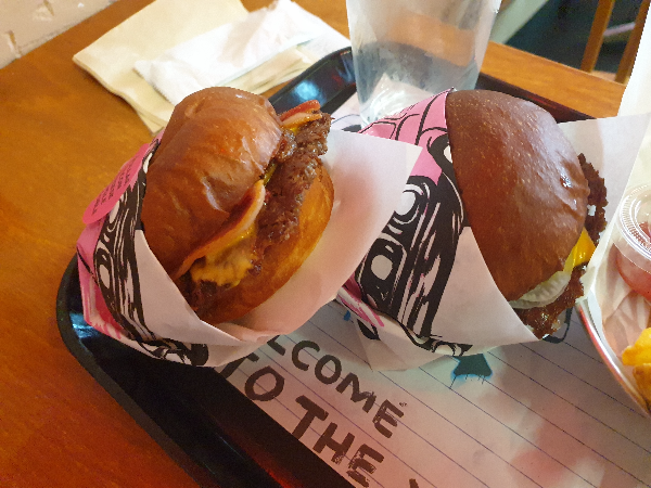 우드 테이블에 쟁반이 놓여있고 쟁반에는 치즈 버거와 베이컨 버거가 놓여있다.