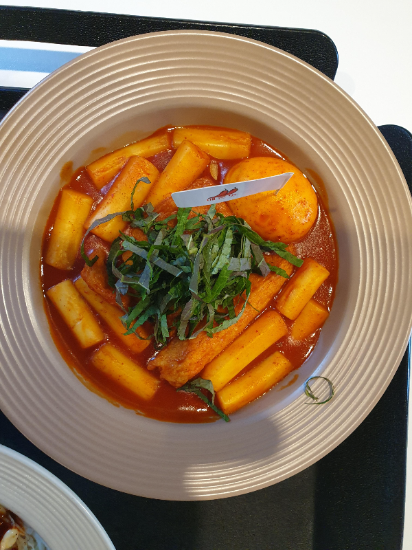 베이지색의 파스타 접시에 나온 밀 떡볶이. 깻잎과 계란 그리고 떡으로 구성되어 있다.