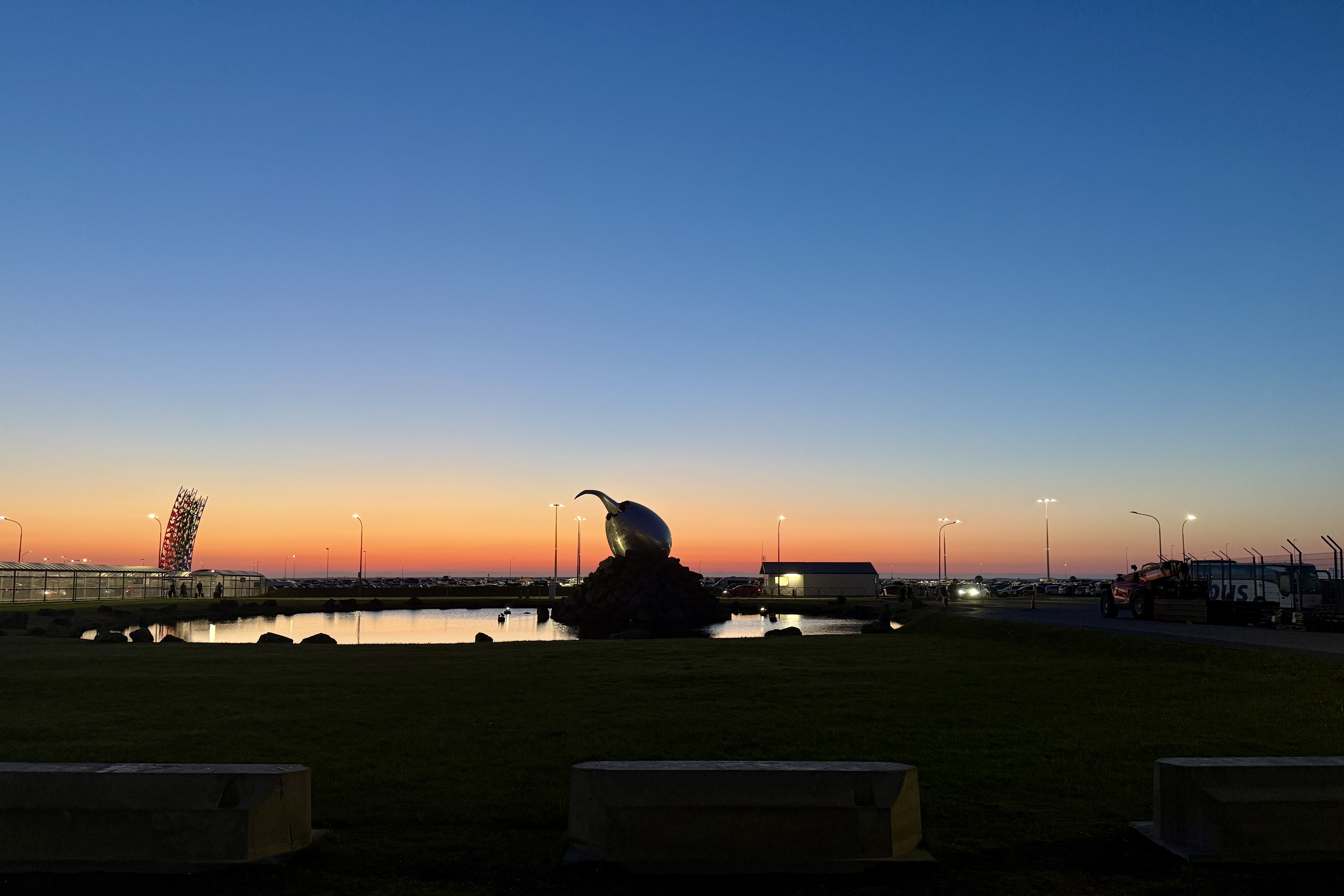 아이슬란드 케플라비크 공항 외부의 백야 풍경. 해가 지지 않은 붉은 하늘 아래 조형물과 반사되는 호수, 조명이 켜진 공항 주차장이 함께 어우러져 있음.