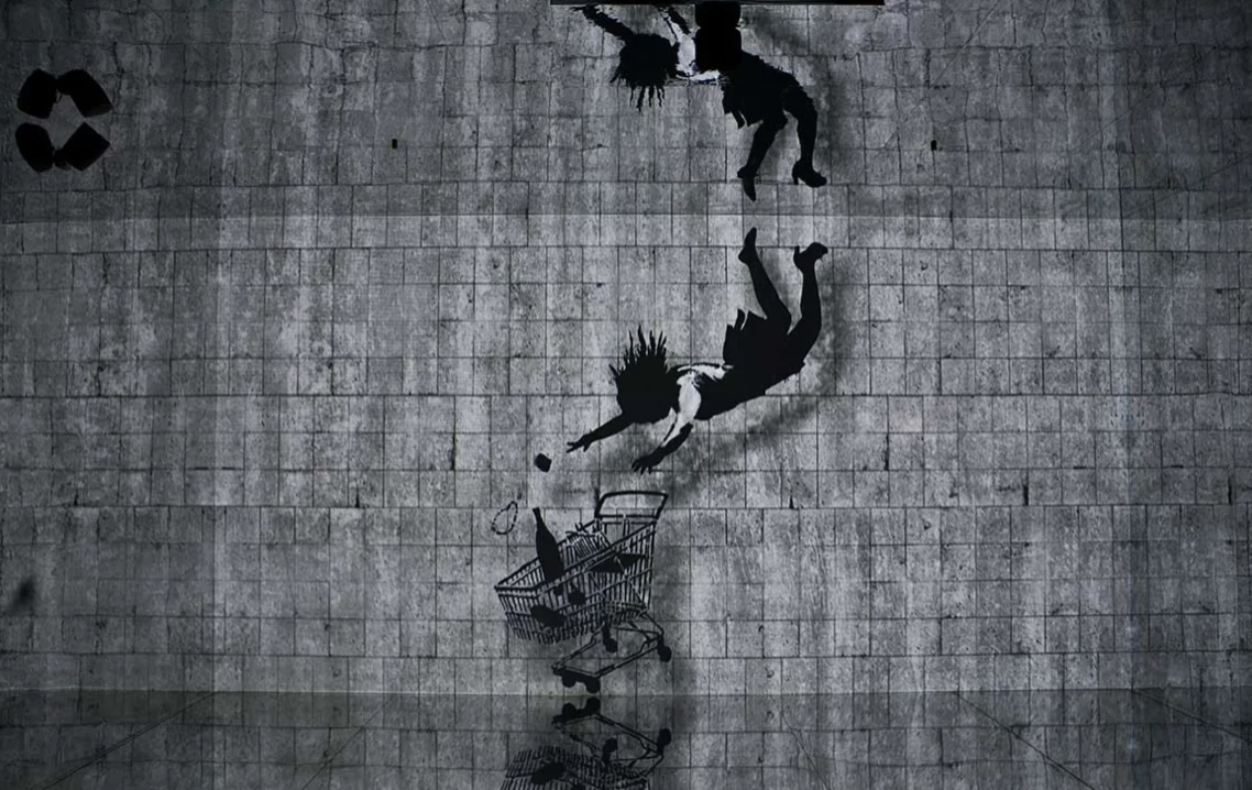 회색 타일 벽면에 검은색 사람 그림이 그려져 있으며, 한 인물이 공중에서 쇼핑카트를 향해 떨어지는 모습이 표현된 그래피티 작품.