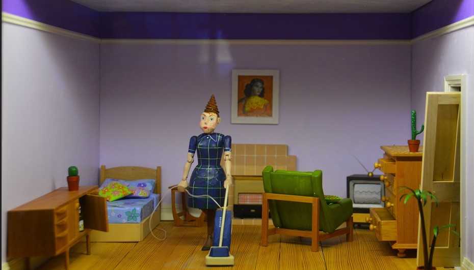 그림이 걸려있고, 침대와 소파 등 가구가 있는 방안에 꼭두각시 인형이 진공청소기를 들고 있는 그림