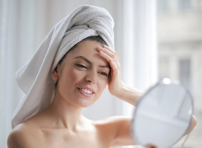 수건을 두른 여성이 거울을 보며 피부를 점검하는 모습으로, 피부타입별 맞춤 스킨케어 루틴의 필요성을 시각적으로 표현합니다.